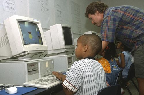 Students working on old computers while a teacher assists